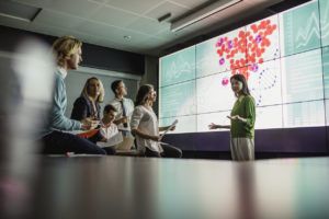 Group of biostatistics professionals in a dark room standing in front of a large data display screen with information.